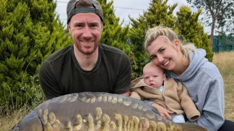 Handout Mum with blonde hair cuddling her baby who is wearing a brown jacket. They are stood next to a man in a green baseball cap holding a big fish in a field. 