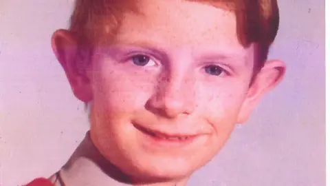 Brian McDermott in a school photo. The image is slightly worn but the young boy is smiling and has short ginger hair and blue eyes and some freckles on his face. 