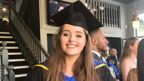 Grace Millane pictured in front of a grand staircase at her university graduation. She is smiling.