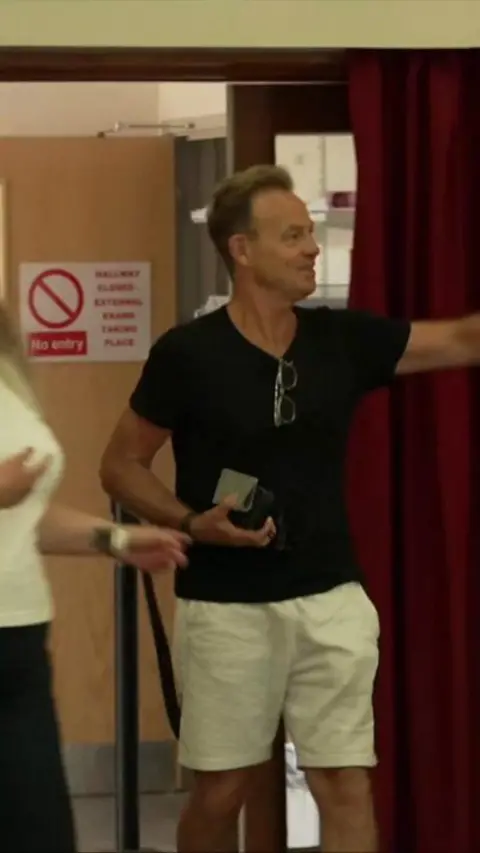 A man with a black t-shirt and shorts on pointing to the side of the frame.