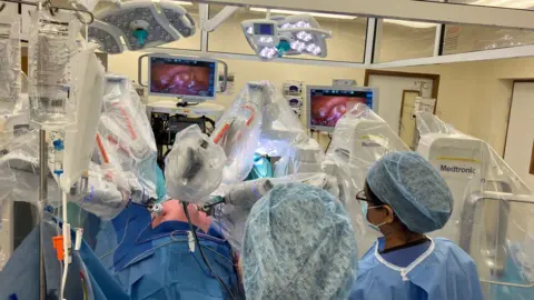 East Kent Hospitals University NHS Foundation Trust Hospital staff and machinery pictured, alongside a patient undergoing a robot-assisted hysterectomy.