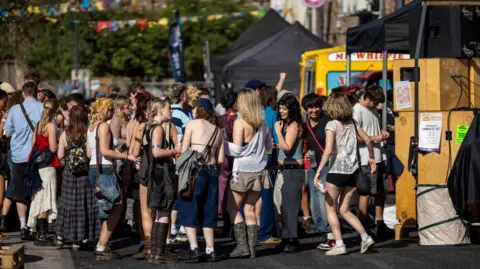 Jam on the Horizon A street party outside the Jam Jar, in St Jude's, Bristol. There are around 30 people in shot, wearing party clothes of skirts, shorts, dresses, t shirts, and bags. Many of them are smiling and looking happy, chatting with other people who are at the party.
There is a Mr Whippy ice cream van to the right of the picture and party flags to the left. It looks like a sunny day.
