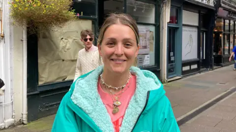 Tiffany is smiling at the camera. She has blonde hair and is wearing a pink t-shirt and a green fleece.