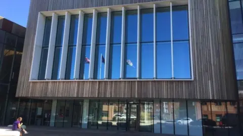 The civic centre in Redcar is square and has a large window. The window appears to be blue and flags are reflected in the glass. The ground floor is glass fronted and cars are reflected in the panes. There is a woman sitting on a white stone seat outside to the left. 