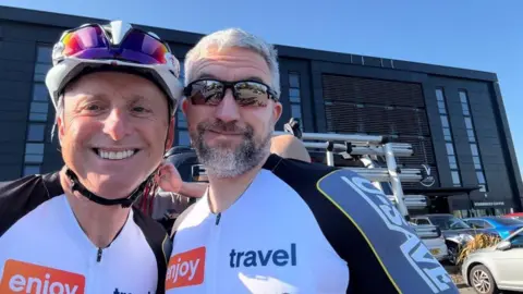 Andy North and Martin Mansell in cycling gear, standing together in front of a building which is covered in modern black cladding. 