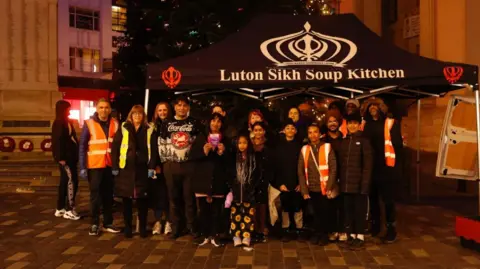 Kian Boyle/BBC A large group of adults and children stand together at night beneath a black canopy labeled “Luton Sikh Soup Kitchen". They face the camera, some wearing hi-vis vests, with a lit Christmas tree and surrounding buildings behind them.