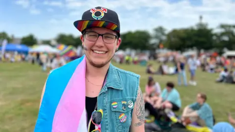 Sam stands in a park during an event. He wears a black cap with a multicoloured gender symbol, a denim vest with various pins and patches, and sunglasses hanging from their shirt. A transgender pride flag is draped over his shoulder. In the background, people are sitting on the grass near tents and booths under a partly cloudy sky.