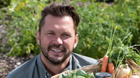Jimmy Doherty has long brown hair swept back. He is smiling while squinting slightly and holding a wicker basket full of fruit and vegetables.