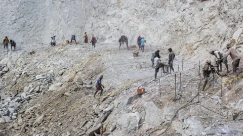 Mukesh Ka Landscape shot for a vast quarry showing many workers working hard to dig work, with yellow energy cables climbing over the ground