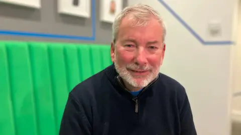 BBC A man sits in an office building on a green bench seat. There are pictures on the wall behind him. 