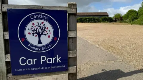 Andrew Turner/BBC Cantley Primary School car park sign - bearing those exact words, stating "users of this car park do so at their own risk". The logo features a blue tree, with blue and some red leaves. In the backdrop are the school buildings, made from timber, and the school car park.