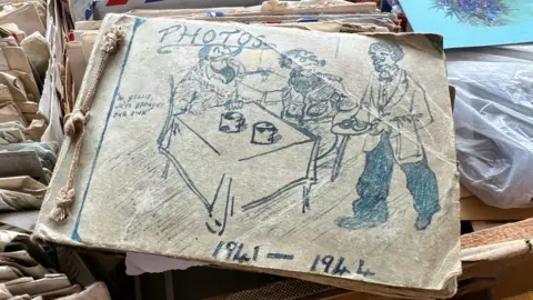 Mike Gould A very old and worn photo album with rope binding. We are looking at its cover, which has a cartoon sketch on the front showing a man bringing tea to a table where two other people are sitting. On the cover it says photos 1941 to 1944. There is also a quote which says "No grazie, we've brought our own".