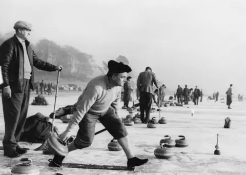 Getty Images A black and white picture of a man in a bunnet and plus fours tossing a curling stone across a frozen loch