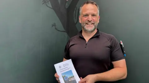 Image of by Insp Andrew Miller wearing a black short sleeved shirt, standing in front of a portion of the mural which features the base of a tree. He is holding a copy of the Custody Handbook.