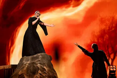 AFP Greek singer Klavdia Papadopoulou, known as Klavdia representing Greece at Eurovision 2025, sings with her arm outstretched on top of a rock stage prop with a fiery background.