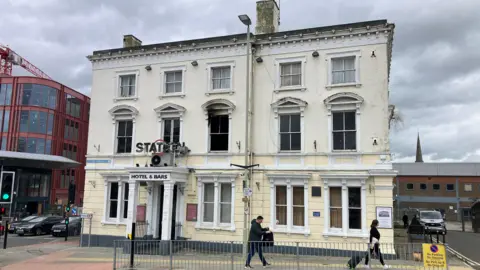 An exterior view of the Station Hotel. It is an older building and rendered with cream and white plaster. Some of the windows on the first floor appear to be missing glass and one of the windows has a black scorch mark above it. 