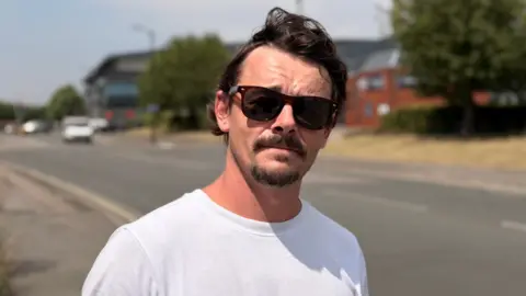A man wearing sunglasses and a white tshirt stood on a trading estate looking into the camera 
