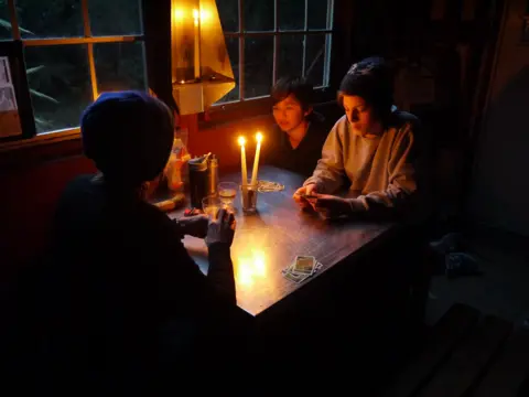 Tony Dowell Three people playing cards at a table by candlelight. They are sitting beside a window and it is dark outside.