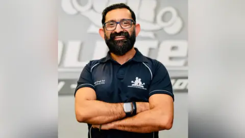 Health Scientist & Body Transformation Coach, Manjit Singh Dol, with dark hair, a beard and glasses smiling with his arms folded and wearing a black t-shirt against a faded grey background. 