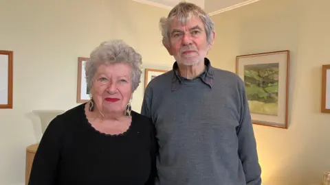 A woman with short grey hair and large earrings wearing a black top, stood next to her husband who is taller than her, wearing a shirt and grey jumper and with grey hair. 