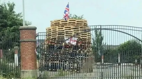 Archive image of the Adam Street bonfire in Belfast. There are bonfire pallets piled up and a white flag with a red cross on it. There is a gate in front of it.