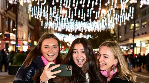 PA Media Three girls take a photo of themselves by Oxford Street lights