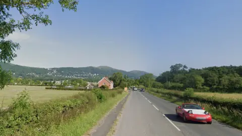 The straight road stretches out into the countryside with hills in the background. There is a house on the left hand side and cars driving on either side.