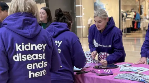 Kidney Disease UK A stall in Queensgate Shopping Centre which is covered in pink fabric and has piles of leaflets fanned out on its top. A woman in a purple hoody saying Kidney Research UK is leaning across the table to another couple of women, facing her in purple hoodies which say on their backs kidney disease ends here. 