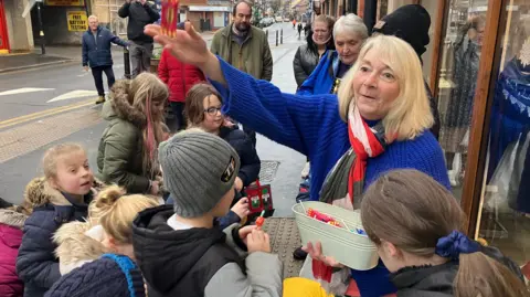 A woman with shoulder-length blonde hair wearing a blue jumper and scarf mid-action throwing a sweet in the air with a crowd of children around her waiting to catch. They are on a street corner outside a shop.