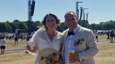 Kim Gladman Kim and Gareth Gladman pose together near the Oasis stage before the concert in their wedding outfits. Fans can be seen milling around in the background.