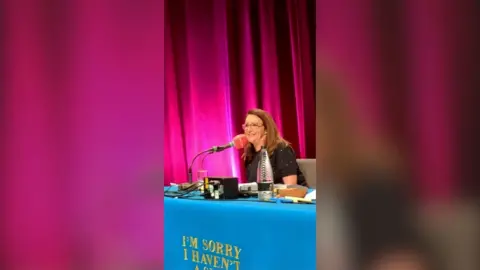 A woman sits behind a table and speaks into a microphone. She has long brown hair and glasses.