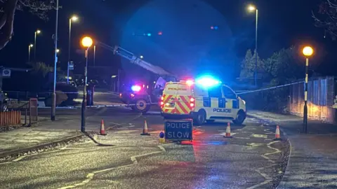 Supplied A police cordon in Chitterman Way in Markfield, Leicestershire. There is a telehandler in the middle of the road outside a shop with a police van parked in front of it, with blue lights on. 