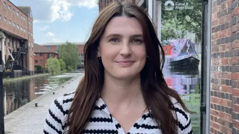 Rachel is standing by the canal wearing a black and white stripy top . She has long brown hair.