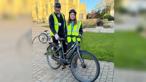 Gloucestershire County Council Councillors David Gray and Chloe Turner standing in front of Gloucester cathedral with an e-bike and wearing bicycle helmets and hi-vis jackets.