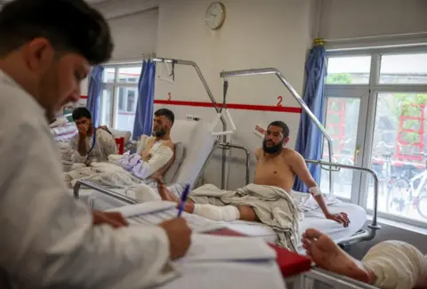 A doctor takes notes at a hospital in Kabul, where three men sit in hospital beds. Two of them have bandages - one to a leg, and another to his arm. The bandaged leg of a fourth men can be seen in the side of the shot.