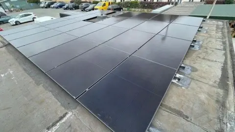 James Middleton A view of black solar panels on top of a building. The roof underneath is grey. On the ground level to the left there is a car park with parked cars.