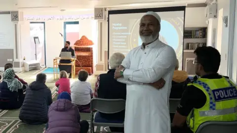 A man dressed in white stands in a mosque smiling towards the camera. Next to him, a police officer is sitting among a group of people listening to a presentation about forthcoming programmes at the mosque.