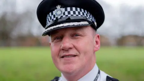 Image of a man in police uniform smiling at the camera. In the background can be seen green fields and trees.