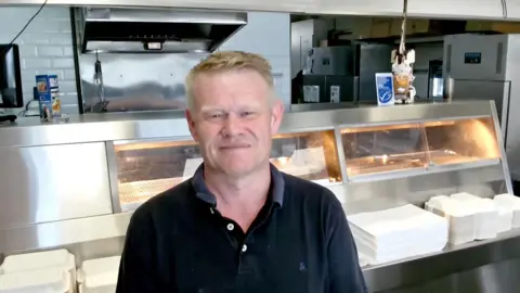 Andrew stands in front of a fish frier in a shop. He's smiling and wearing a dark polo top. 