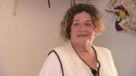 Lucy Bennett looking to the left and wearing a white shirt and wool gilet. She has short brown hair and is stood in her son's flat which has white walls.