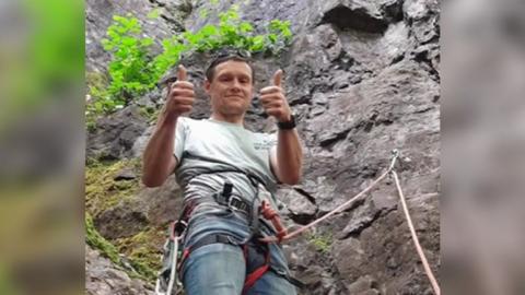 A man stands on a rocky outcrop with a steep cliff behind him. He's wearing a climbing harness attached loosely to the rock behind and is giving a thumbs up and smiling.