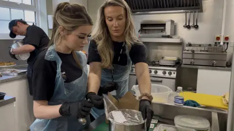 A woman with long blonde hair is helping a teenage girl removing clingfilm from a large put on a counter.
The pair are wearing blue aprons and black latex gloves,
There is man in the backgroun
