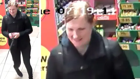 A screenshot from CCTV inside a shop of a woman wearing black trousers, a black jacket and a black rucksack. She has dark blonde hair tied back.