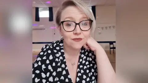 Ellie Howells pictured wearing a black dress with white hearts. Ellie has blonde hair and black-rimmed glasses.