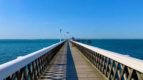 Jungle Jim A pier going out into the water with the dark blue ocean either side and clear blue skies above. There is a British flag flying at the end of the wooden pier.