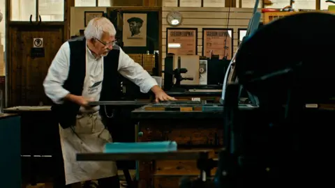 A man with white hair and glasses. He is wearing a white shift with a black sleeveless jacket and an apron around his waist. He is pulling a metal bar away from a table. He is focussed on the table to his left. In the background, the walls are busy with artwork. To the far left in the foreground, there is the silhouette of machinery. 