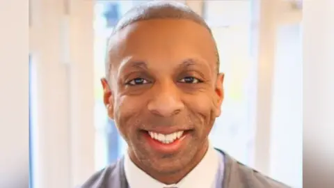 Martyn Latchman: a professional head‑and‑shoulders portrait of a man indoors. He is wearing a light shirt under a grey jacket, and looking directly at the camera with a smiling expression.