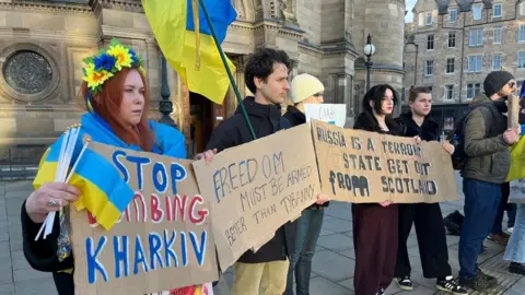 A row of people with banners relating to the war in Ukraine. A woman nearest the camera is wearing Ukrainian national dress with a flower garland in her hair and is carrying a number of small light blue and yellow Ukrainian flags. A man next to her has a large Ukrainian flag.