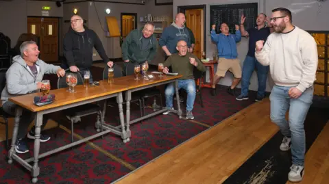 Joss Barratt A group of men are cheering in a skittle alley after performing well at skittles. Two men are sitting at two long tables which have drinks on them.