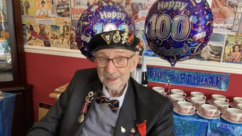 BBC Dennis Lanham stood in front of some "Happy 100th Birthday" balloons and a table full of teacups ready for a party
